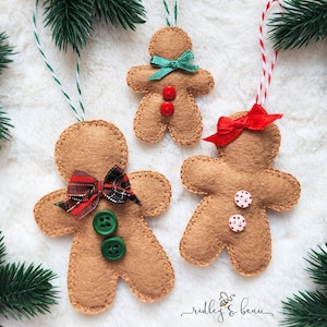 May include: Three tan felt gingerbread person ornaments with stitched edges. One has a plaid bow and green buttons, one has a red bow and white polka dot buttons, and one has a green ribbon bow and red buttons, all with hanging strings.