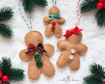 May include: Three tan felt gingerbread person ornaments with stitched edges. One has a plaid bow and green buttons, one has a red bow and white polka dot buttons, and one has a green ribbon bow and red buttons, all with hanging strings.