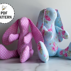May include: Two handmade plush bunny toys. One is pink with white polka dots and the other is blue with a floral pattern. Both bunnies have long ears and are sitting on a white surface.