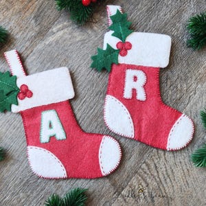 May include: Two red felt Christmas stockings with white cuffs and toes. Each stocking features a green felt holly leaf and red berry accent on the cuff. One stocking has a white letter A and the other has a white letter R stitched on the front.
