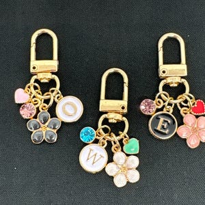 Op de afbeelding: Drie gouden sleutelhangers met bedels. Elke sleutelhanger heeft een letterbedel, een bloemenbedel, een hartbedel en een kristalbedel. De letters zijn O, E en W.