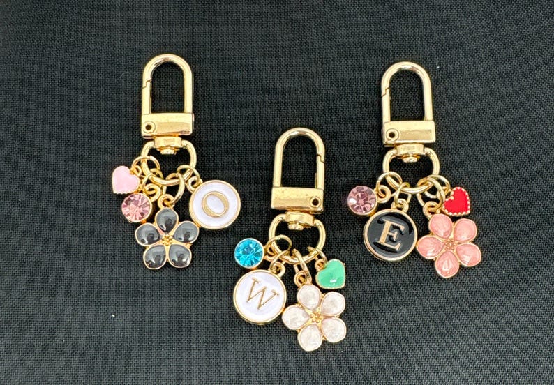 Op de afbeelding: Drie gouden sleutelhangers met bedels. Elke sleutelhanger heeft een letterbedel, een bloemenbedel, een hartbedel en een kristalbedel. De letters zijn O, E en W.