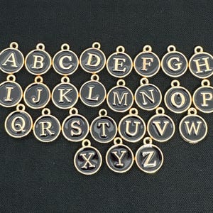 Op de afbeelding: Een set van 26 gouden metalen alfabetbedels met zwart email. Elke bedel heeft een letter van het alfabet in een typemachinelettertype.