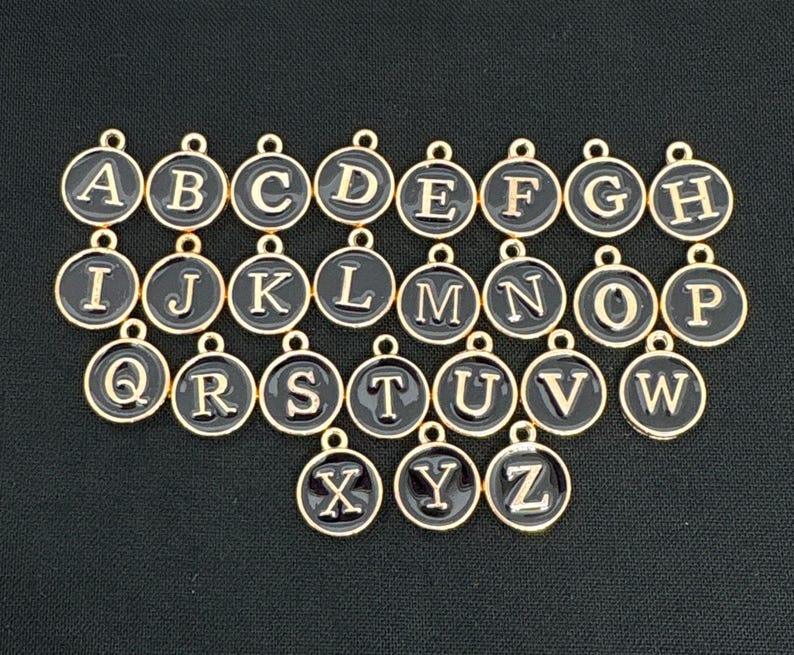 Op de afbeelding: Een set van 26 gouden metalen alfabetbedels met zwart email. Elke bedel heeft een letter van het alfabet in een typemachinelettertype.
