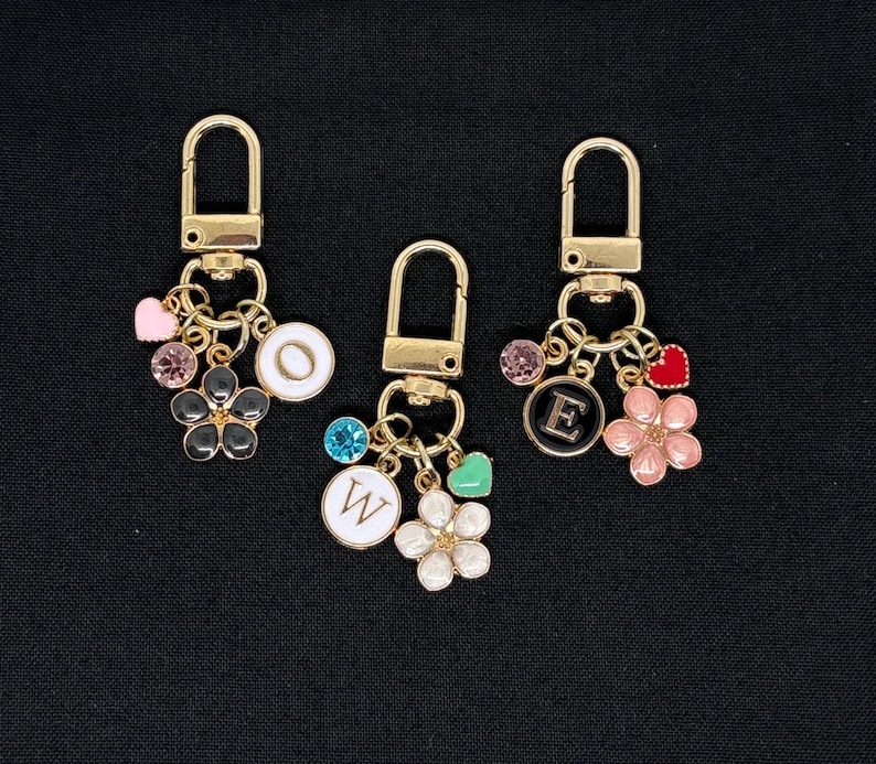 Op de afbeelding: Drie gouden sleutelhangers met bedels. Elke sleutelhanger heeft een letterbedel, een bloemenbedel en een hartbedel. De letters zijn "O", "E" en "W".