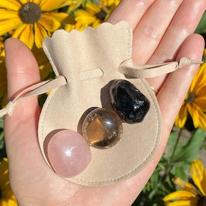 May include: A small, beige suede pouch with a drawstring closure holds three tumbled stones: a pink rose quartz, a clear smoky quartz, and a black obsidian.