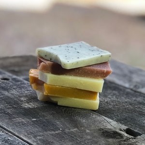Puede incluir: Una pila de barras de jabón hechas a mano en varios colores, incluyendo verde claro con motas oscuras, marrón rojizo, amarillo y naranja. Los jabones son cuadrados y están apilados sobre una superficie de madera envejecida, mostrando una estética natural.