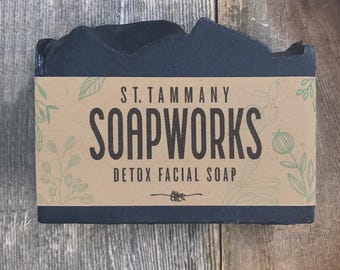 Puede incluir: Una pastilla de jabón facial desintoxicante de carbón negro con una etiqueta de papel marrón que dice "St. Tammany Soapworks Detox Facial Soap".