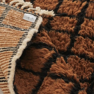 May include: A close-up of a brown and black textured rug. The rug has a geometric pattern with a shaggy texture. The edge of the rug is visible, showing a striped pattern in tan and black.