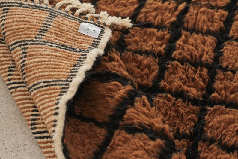 May include: A close-up of a brown and black textured rug. The rug has a geometric pattern with a shaggy texture. The edge of the rug is visible, showing a striped pattern in tan and black.