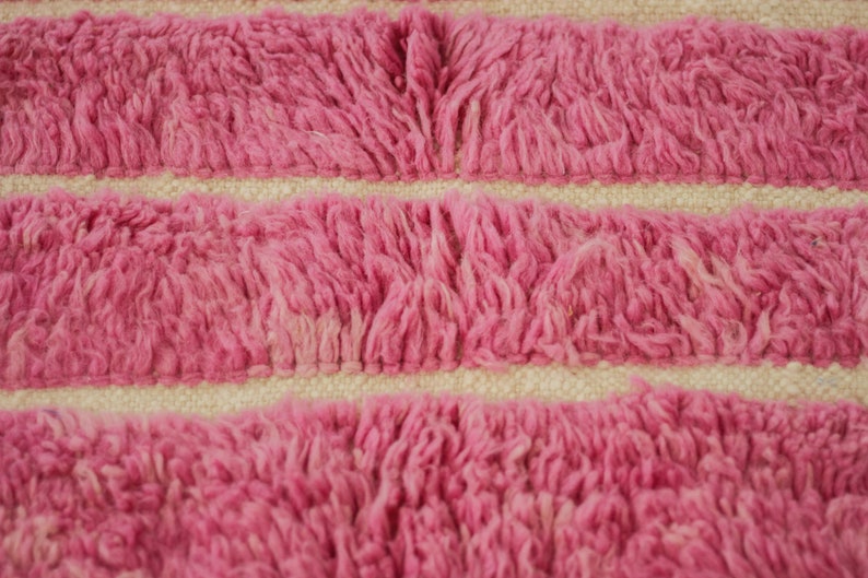 May include: Close-up of a pink and white striped rug with a fuzzy texture. The rug is made of woven fibers and has a soft, plush feel.