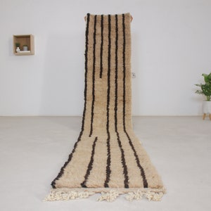 Op de afbeelding: Een lang, smal beige kleed met donkerbruine strepen. Het kleed heeft franjes aan de uiteinden.