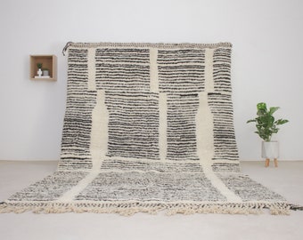 Puede incluir: Una alfombra rectangular grande con un patrón de rayas blancas y negras. La alfombra presenta gruesas rayas negras horizontales con rayas blancas verticales. La alfombra tiene flecos en el borde inferior y se muestra contra una pared blanca.