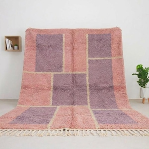 May include: A rectangular area rug featuring a shaggy texture with a geometric pattern of large purple blocks set against a dusty pink background. The rug is finished with cream-colored fringe along the bottom edge, creating a soft textile surface.