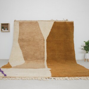 May include: A hand-woven rug with a geometric design in shades of beige, brown, and white. The rug has a fringe border and a subtle purple stripe.