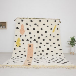 May include: A white rug with black polka dots and abstract shapes in yellow, pink, and black. The rug has a fringe border.