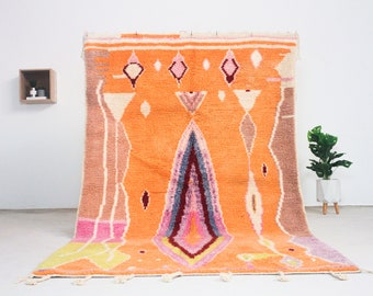Puede incluir: Una alfombra grande tejida a mano con un patrón geométrico en tonos naranja, rosa, morado y marrón. La alfombra tiene un borde de flecos y está colgada en una pared blanca.