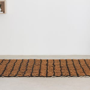 May include: Rectangular brown and black rug with a geometric pattern. The rug has a textured surface and a cream-colored fringe on the short ends. The rug is placed on a light-colored floor.