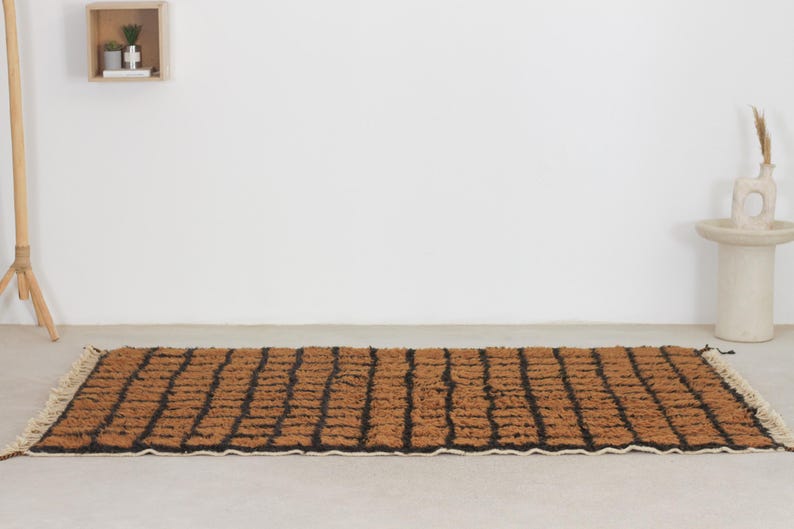 May include: Rectangular brown and black rug with a geometric pattern. The rug has a textured surface and a cream-colored fringe on the short ends. The rug is placed on a light-colored floor.
