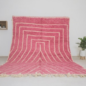 May include: A pink and white striped Moroccan rug with a geometric pattern. The rug has a fringe border and is made of wool.