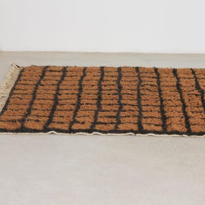 May include: A rectangular brown rug with a black grid pattern. The rug has a textured, woven appearance and a cream-colored fringe along one edge. The rug is placed on a light-colored surface.