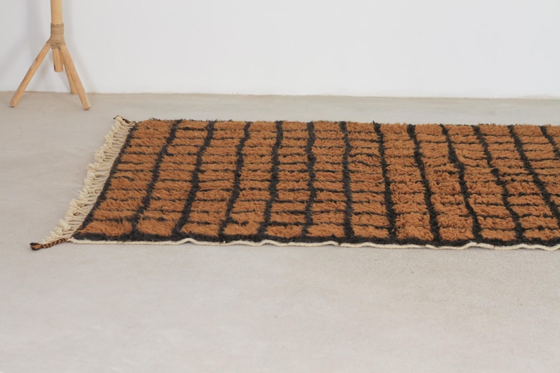 May include: A rectangular brown rug with a black grid pattern. The rug has a textured, woven appearance and a cream-colored fringe along one edge. The rug is placed on a light-colored surface.