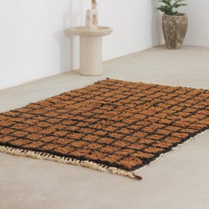 May include: A rectangular brown rug with a black grid pattern. The rug has a textured surface and a fringe along the edges. The rug is placed on a light-colored floor.