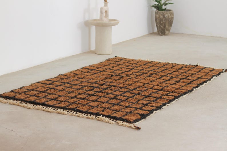 May include: A rectangular brown rug with a black grid pattern. The rug has a textured surface and a fringe along the edges. The rug is placed on a light-colored floor.