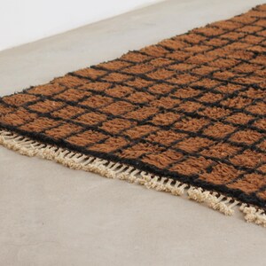 May include: A rectangular brown rug with a black grid pattern. The rug has a shaggy texture and a cream-colored fringe along one edge. The rug is placed on a light gray surface.