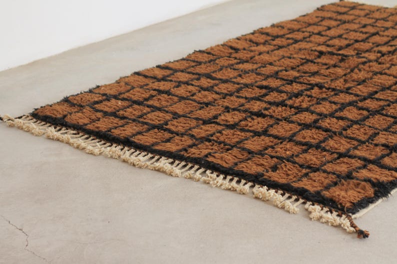 May include: A rectangular brown rug with a black grid pattern. The rug has a shaggy texture and a cream-colored fringe along one edge. The rug is placed on a light gray surface.