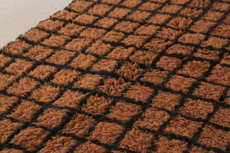 May include: Close-up of a brown, textured fabric with a repeating square pattern. The squares are outlined in black, creating a grid-like design. The fabric has a soft, plush appearance.
