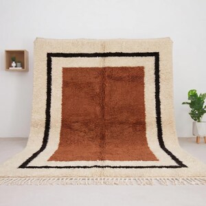 May include: A large, rectangular rug with a cream-colored background and a central brown section. The rug features a black border and fringe along the bottom edge. The rug is displayed against a white wall, suggesting a home decor item.