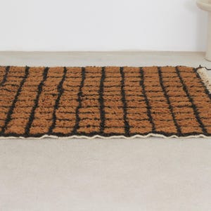 May include: A rectangular brown rug with a black grid pattern. The rug has a textured surface and a white fringe along one edge. The rug is placed on a light gray surface.