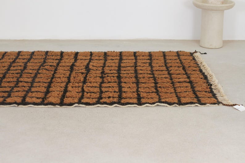 May include: A rectangular brown rug with a black grid pattern. The rug has a textured surface and a white fringe along one edge. The rug is placed on a light gray surface.