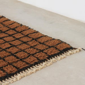May include: A rectangular rug with a brown and black checkered pattern. The rug has a textured, woven appearance with a cream-colored fringe along one edge. The rug is placed on a light gray surface.