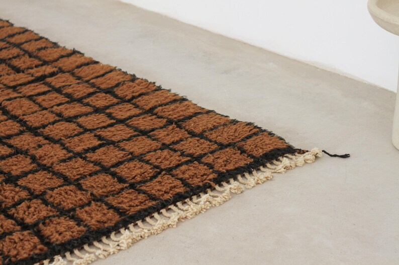 May include: A rectangular rug with a brown and black checkered pattern. The rug has a textured, woven appearance with a cream-colored fringe along one edge. The rug is placed on a light gray surface.