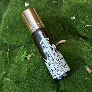 May include: A dark brown glass roller bottle with a gold cap. The bottle has a white design of a sword and mushrooms.