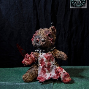 Puede incluir: Un oso de peluche marrón con manchas rojas y un cuchillo ensangrentado en su mano. El oso lleva un delantal blanco con manchas rojas y un collar negro con tachuelas. El oso está sentado sobre una superficie verde.