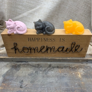 May include: Three handmade soap bars shaped like sleeping cats. The soaps are pink, gray, and yellow. The soaps are sitting on a wooden sign that says "Happiness is homemade".