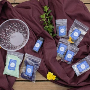 May include: An assortment of items for a craft project, including a clear glass bowl, small bottles, and several small plastic bags filled with colorful materials. The items are arranged on a burgundy fabric.