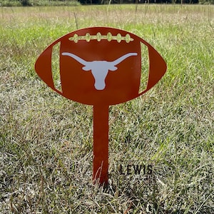 Peut inclure: Un piquet de jardin en m&eacute;tal orange en forme de ballon de football avec un logo de longhorn blanc. Le piquet est plant&eacute; dans un champ herbeux sous un ciel bleu clair. Le ballon a un design d&eacute;coup&eacute;.