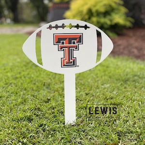 May include: White metal football yard sign with a red and black Texas Tech University logo. The sign is on a green grassy lawn.
