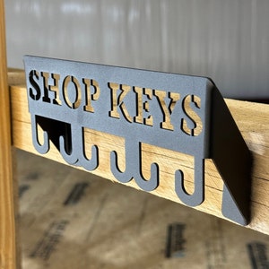 May include: Black metal key rack with the words "SHOP KEYS" cut out and five hooks. The rack is mounted on a wooden surface.