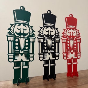 May include: Three nutcracker figurines, one green, one black, and one red, standing on a wooden surface. Each nutcracker has a white face, a black mustache, and a black beard.