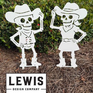 May include: Two white metal skeleton cowboy garden ornaments. One skeleton wears a vest and the other a dress. Both wear cowboy hats and boots. The text "LEWIS DESIGN COMPANY" is at the bottom.
