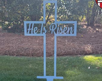 May include: A white metal cross with the words "He Is Risen" cut out in the center. The cross is standing in a grassy lawn.