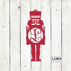 May include: Red metal nutcracker silhouette with a monogrammed circle in the center. The monogram is the letter "L". The nutcracker is standing against a white wood background. The text "LEWIS DESIGN COMPANY" is in the bottom right corner.
