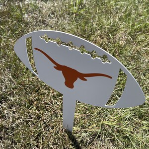 Peut inclure: Un piquet de jardin en m&eacute;tal blanc en forme de ballon de football, orn&eacute; d'un logo longhorn orange br&ucirc;l&eacute;. Le piquet pr&eacute;sente des d&eacute;tails d&eacute;coup&eacute;s et est plant&eacute; dans l'herbe, pour la d&eacute;coration ext&eacute;rieure.