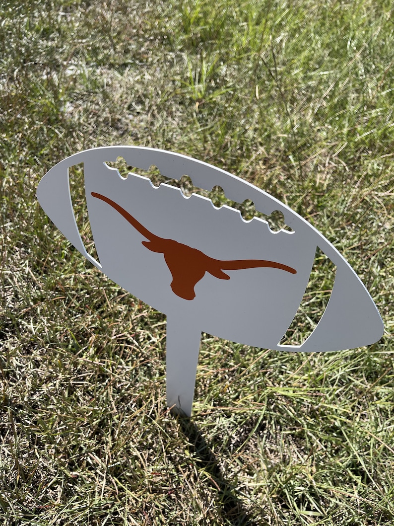 Peut inclure: Un piquet de jardin en m&eacute;tal blanc en forme de ballon de football, orn&eacute; d'un logo longhorn orange br&ucirc;l&eacute;. Le piquet pr&eacute;sente des d&eacute;tails d&eacute;coup&eacute;s et est plant&eacute; dans l'herbe, pour la d&eacute;coration ext&eacute;rieure.