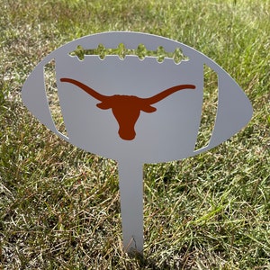 Peut inclure: Un piquet de jardin en m&eacute;tal blanc en forme de ballon de football, orn&eacute; d'un logo de longhorn orange br&ucirc;l&eacute;. Le piquet pr&eacute;sente des d&eacute;tails d&eacute;coup&eacute;s et est plant&eacute; dans l'herbe, id&eacute;al pour la d&eacute;coration ext&eacute;rieure.